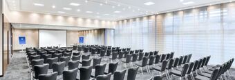 Conference room at NH Hotel M&uuml;nchen Neue Messe with rows of black chairs, projection screen, and roll-up boards