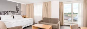 Hotel room at NH Hotel M&uuml;nchen Neue Messe featuring a double bed, a sofa, coffee table, armchair, and balcony doors with curtains