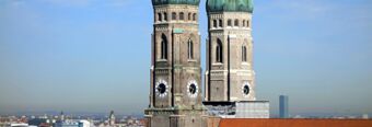 Two tall brick clock towers with green domes overlooking a cityscape under a clear blue sky in Munich near NH Hotel M&uuml;nchen Neue Messe