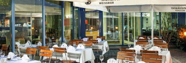 Outdoor dining area with wooden chairs and white tablecloths under large umbrellas at NH Hotel M&uuml;nchen Neue Messe