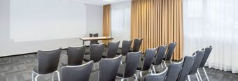 Empty conference room with rows of black chairs, a table with a glass bottle, and beige curtains at NH Hotel M&uuml;nchen Neue Messe