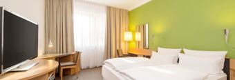 Hotel room at NH Hotel M&uuml;nchen Neue Messe with a double bed, green accent wall, TV, desk, chair, and a large window with curtains