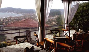 Area ristorante con tavoli e sedie in legno e ampie finestre con tende bianche e vista sulla citt&agrave; presso la Pensione Ristorante Bellavista.