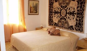 A beige bed with two patterned pillows, two wooden nightstands with lamps, a window with orange curtains, and wall decorations at Pensione Ristorante Bellavista