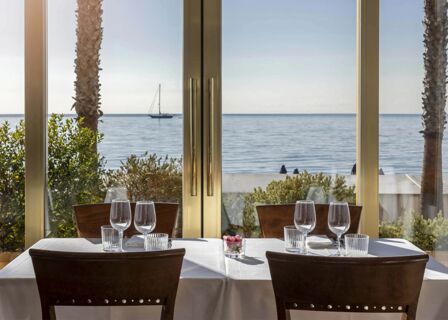 Dining table set with wine glasses and water glasses by large window with sea and sailboat view at B&B Hotel Diano Marina Palace