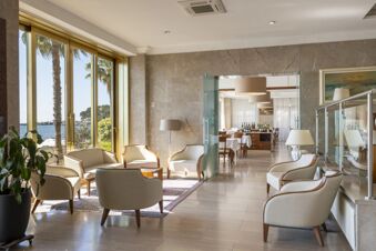 Seating area with cream armchairs and lamps inside B&B Hotel Diano Marina Palace with large glass doors showing palm trees and sea view