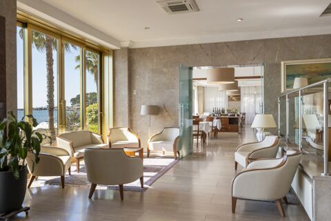 Seating area with cream armchairs and lamps inside B&B Hotel Diano Marina Palace with large glass doors showing palm trees and sea view