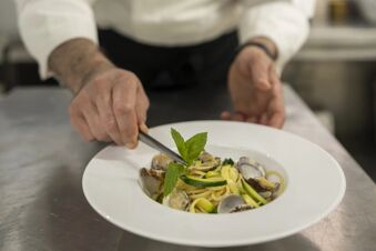 Chef preparing a seafood pasta dish with clams, zucchini, and fresh basil in a white bowl at B&B Hotel Diano Marina Palace