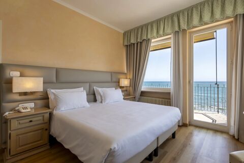 Double bed with white linens and pillows, beige walls, bedside tables with lamps, window and door leading to balcony with sea view at B&B Hotel Diano Marina Palace