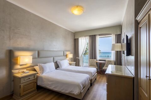 Two single beds with white linens in a hotel room at B&B Hotel Diano Marina Palace featuring a window with sea view and chairs