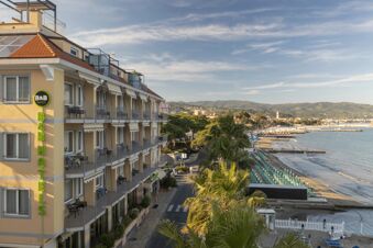 B&B Hotel Diano Marina building with balconies facing a beach lined with umbrellas and a calm sea with mountains in the background