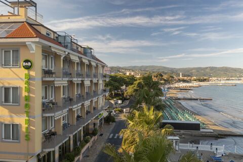 B&B Hotel Diano Marina building with balconies facing a beach lined with umbrellas and a calm sea with mountains in the background
