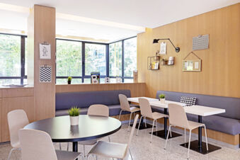 Dining area at B&B Hotel Udine featuring round and rectangular tables with chairs and potted plants near large windows and wooden walls