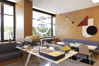 Breakfast buffet with sliced cheese, ham, bread, apples, and seating area at B&B Hotel Udine