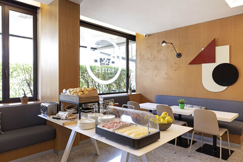 Breakfast buffet with sliced cheese, ham, bread, apples, and seating area at B&B Hotel Udine