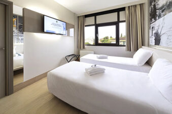 A minimalist twin bedroom at B&B Hotel Udine with two beds, white linens, folded towels, a wall-mounted TV, and a large window