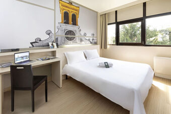 Bright hotel room at B&B Hotel Udine with double bed, white linens, desk with laptop, chair, and large window view of trees