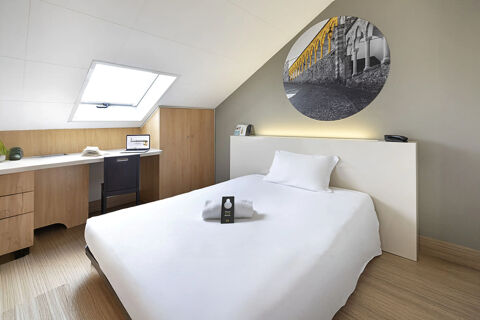 Minimalist hotel room at B&B Hotel Udine featuring a white bed with pillow, wooden desk, chair, and slanted ceiling window