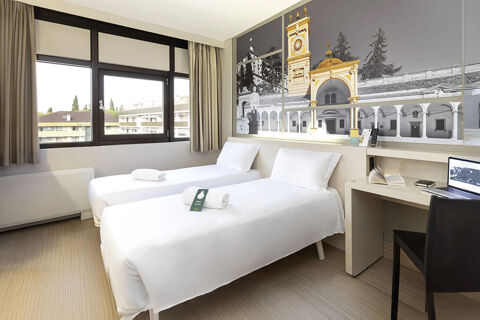Two single beds with white linens and towels in a bright room at B&B Hotel Udine with a large window and desk with a chair
