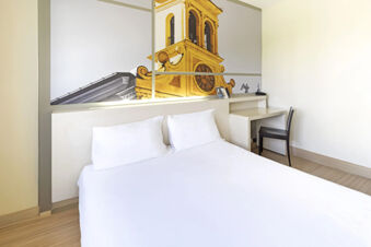Minimalist hotel room at B&B Hotel Udine with a large white bed, two pillows, desk, chair, and wall art of a clock tower