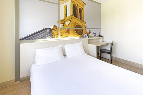 Minimalist hotel room at B&B Hotel Udine with a large white bed, two pillows, desk, chair, and wall art of a clock tower