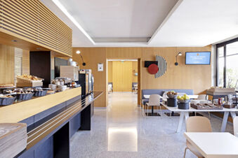 Modern breakfast area at B&B Hotel Udine featuring a wooden buffet with pastries, a seating area, and natural light from windows