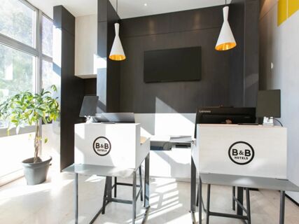 Bright reception area with two white desks branded B&B Hotel Savona, hanging lights, large window, and a potted plant