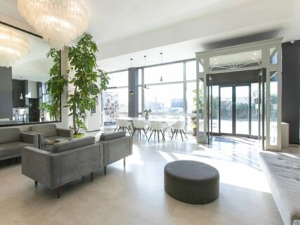 Bright hotel lobby with grey sofas, a round ottoman, dining area by large windows, and plants at B&B Hotel Savona