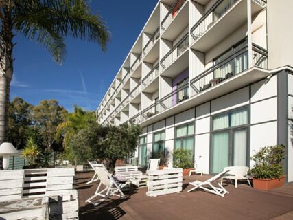 Outdoor wooden terrace with white lounge chairs and tables beside the modern three-story B&B Hotel Savona under a clear sky