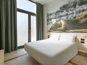 Minimalist hotel room at B&B Hotel Savona with a large bed, white linens, floor-to-ceiling window, and a wall mural of a reclining statue