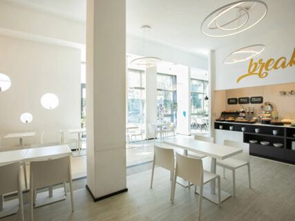 Bright and modern breakfast area at B&B Hotel Savona with white furniture, wooden floor, and a buffet counter on the right side