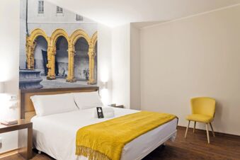 Bright hotel room with white bed that has a yellow throw, yellow chair, side tables, and arch mural at B&B Hotel Palermo Quattro Canti