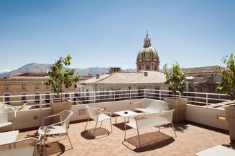 Terrace with white chairs and tables, potted plants, rooftop buildings, mountains, and a dome in the background at B&B Hotel Palermo Quattro Canti