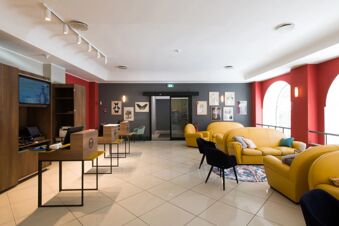 Reception area of B&B Hotel Palermo Quattro Canti with wooden desks, yellow sofas, navy chairs, and botanical wall art