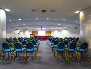 Rows of blue cushioned chairs arranged facing a red table and projector screen in a conference room at B&B Hotel Trapani Crystal