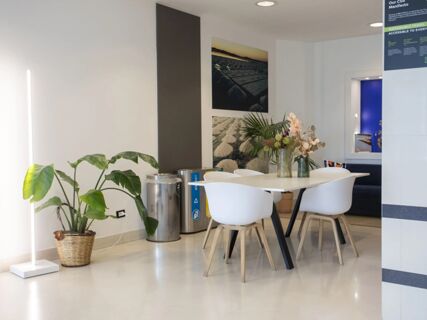 Dining area with white chairs and table, vases with flowers, large potted plant, trash bins, and wall art at B&B Hotel Trapani Crystal