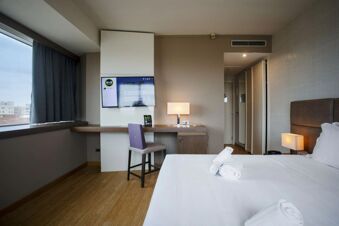Hotel room at B&B Hotel Trapani Crystal with a large bed, rolled towels, a mounted flat-screen TV, desk, chair, and window with curtains