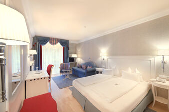 Hotel Ansitz Tschindlhof room with a large bed, blue sofa, striped armchair, desk, and large window with curtains