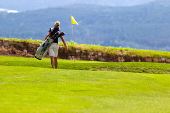 A woman carrying golf clubs walks on a green fairway with a yellow flag near a stone wall at Hotel Ansitz Tschindlhof