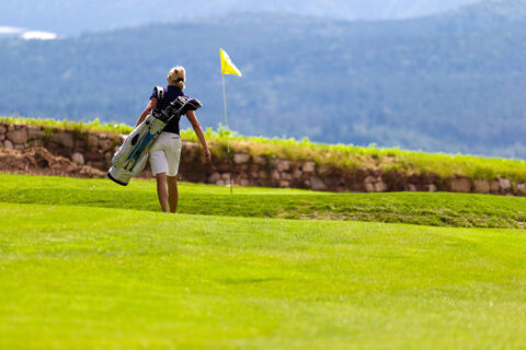 A woman carrying golf clubs walks on a green fairway with a yellow flag near a stone wall at Hotel Ansitz Tschindlhof