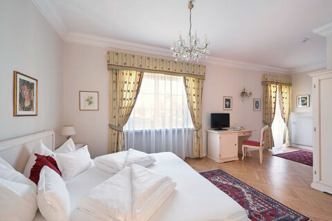 Bright hotel room at Hotel Ansitz Tschindlhof with a white bed, red cushions, wooden floor, desk, chair, and patterned curtains