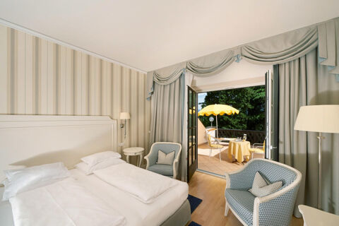 Hotel Ansitz Tschindlhof bedroom with white bed, patterned armchairs, striped wallpaper, and balcony with yellow umbrella and chairs