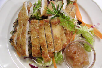 Sliced breaded chicken on mixed greens with carrot slices and herbs, served with a cup of apple sauce at Hotel Ansitz Tschindlhof