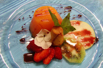 Orange gelatin dessert with sliced strawberry, kiwi, apple, a small cake, mint leaves, and chocolate drizzle on a clear plate served at Hotel Ansitz Tschindlhof