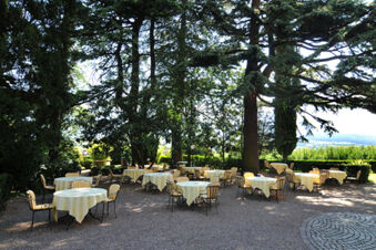 Outdoor seating area at Hotel Ansitz Tschindlhof with multiple round tables covered in white cloths surrounded by trees and sunlight