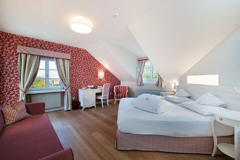 Hotel Ansitz Tschindlhof room with slanted ceiling, double bed with white linens, desk, armchair, sofa, and two windows with curtains