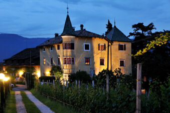 Hotel Ansitz Tschindlhof illuminated at dusk with its yellow facade, stone roof, adjacent vineyard, and mountainous background