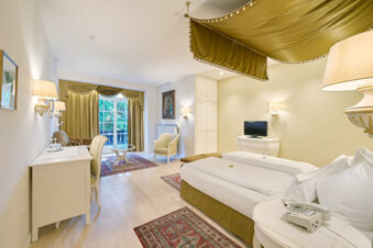 Hotel Ansitz Tschindlhof room with a large bed, desk, armchair, TV on a dresser, and windows with olive green curtains