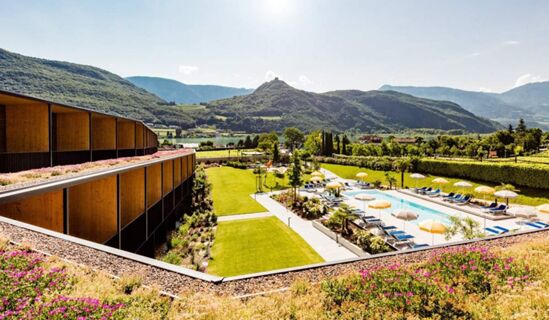 Hotel THALHOF am See features a modern building with balconies, a green lawn, outdoor pool with yellow umbrellas, and surrounding mountains