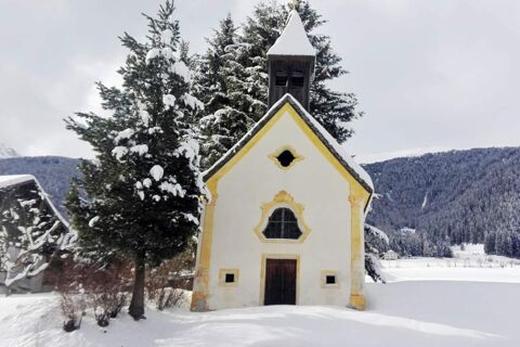 Piccola cappella innevata con decorazioni gialle in inverno presso il maso Innersieslhof, Villa Anna, circondata da abeti e montagne.